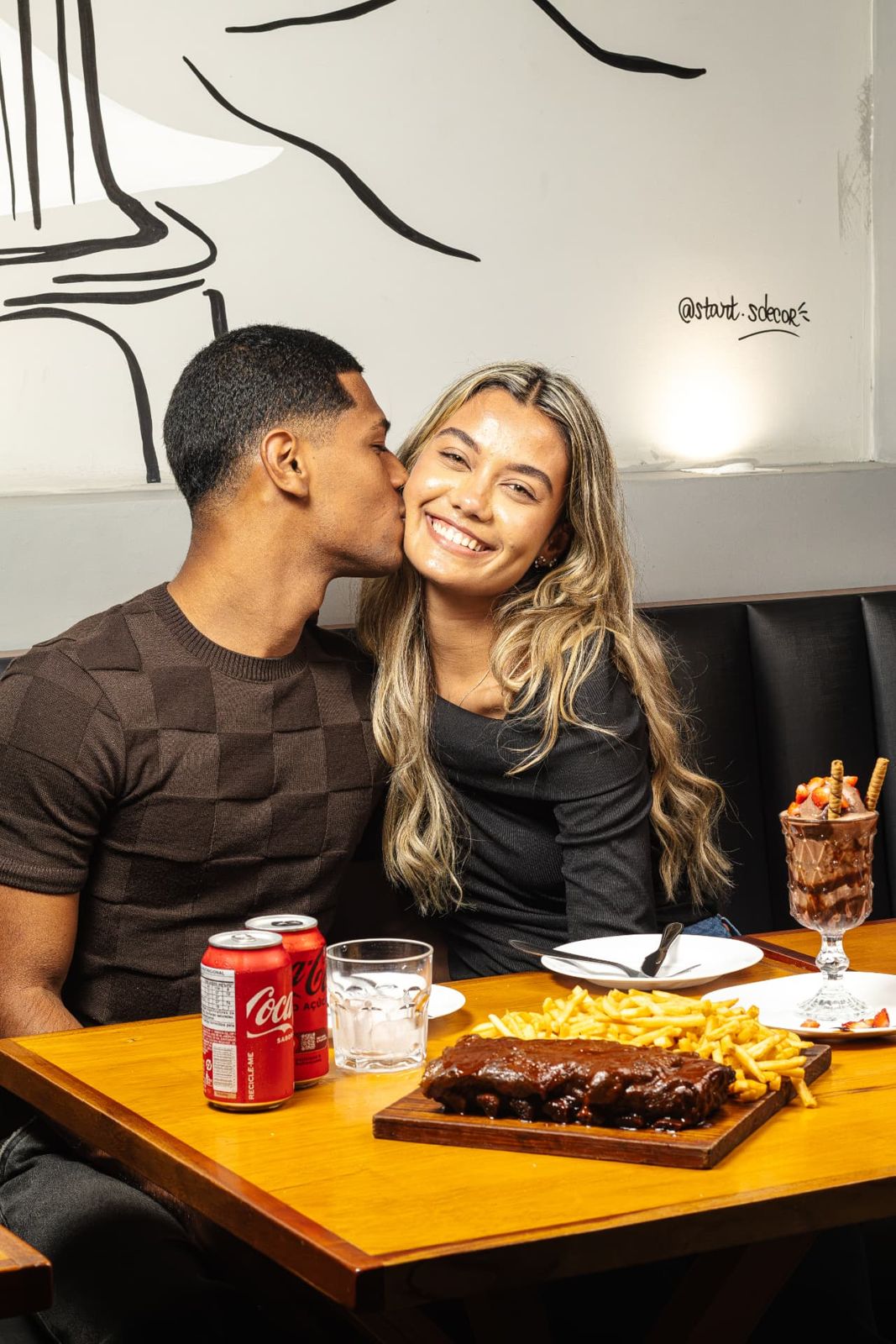 Casal sorrindo à mesa com costela, fritas e sobremesa
